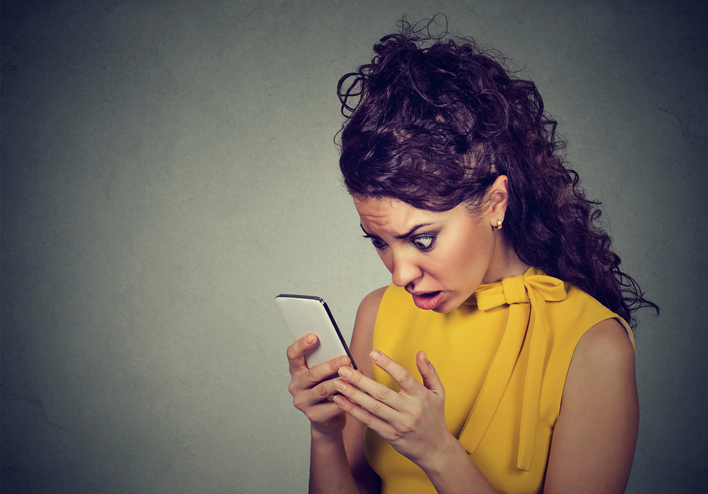 Scared shocked young woman holding cellphone in hands looking at screen with cross face expression at stressful texts and calls isolated on gray background. Negative human emotion